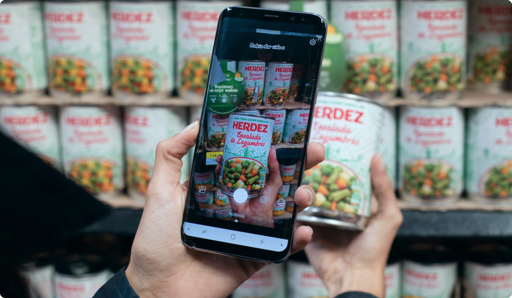 Teléfono móvil escaneando una lata de verduras Herdez en un anaquel de supermercado.